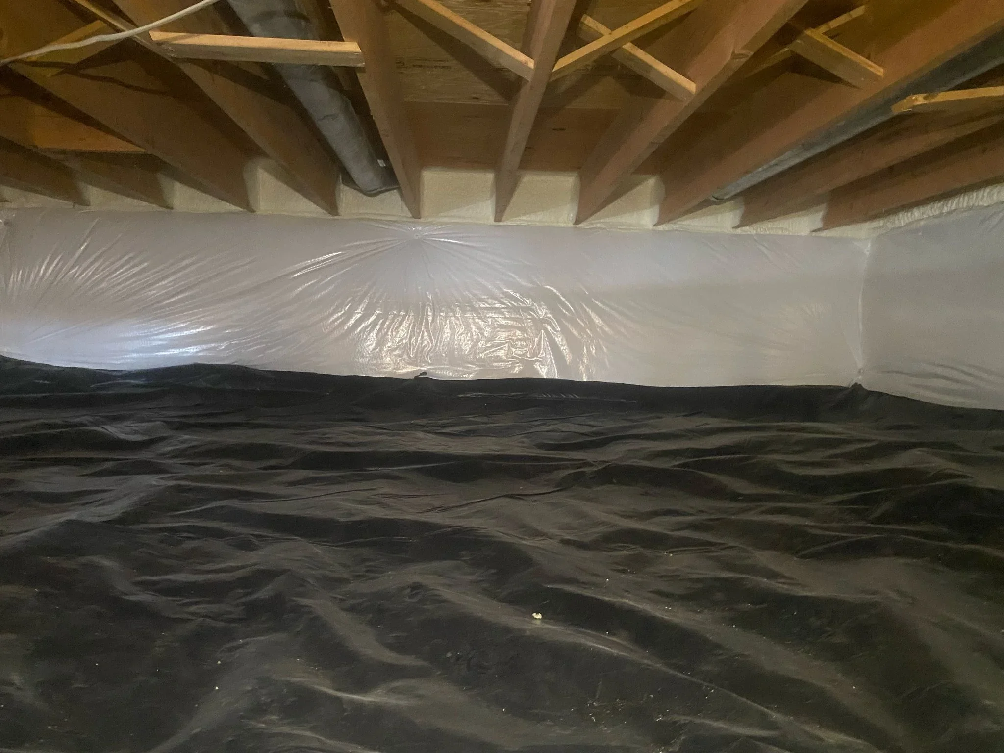 Crawl space with white polyethylene vapor barrier and black ground cover under wooden floor joists