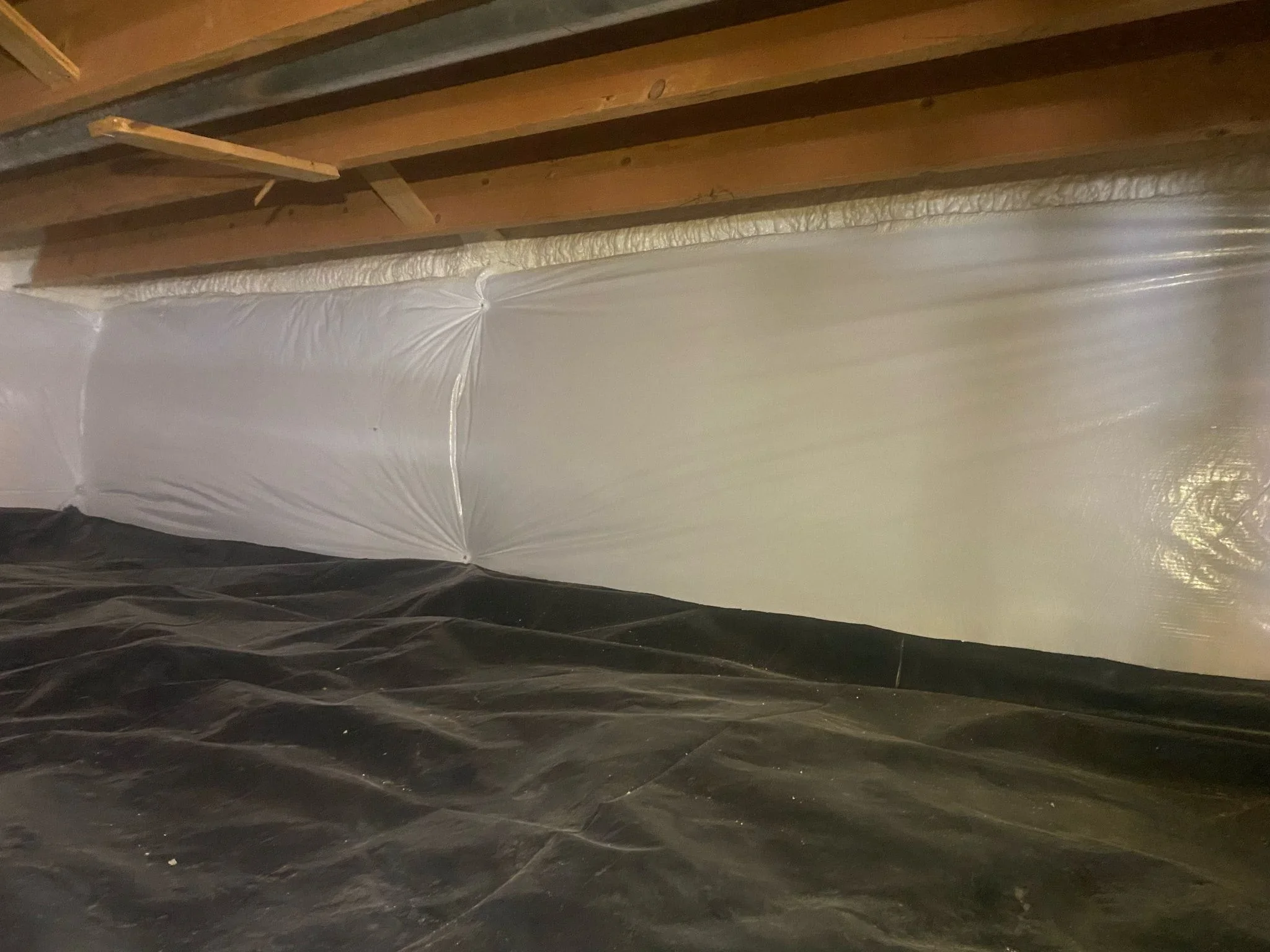 White polyethylene vapor barrier installed in crawl space with black ground cover and wood beam framing